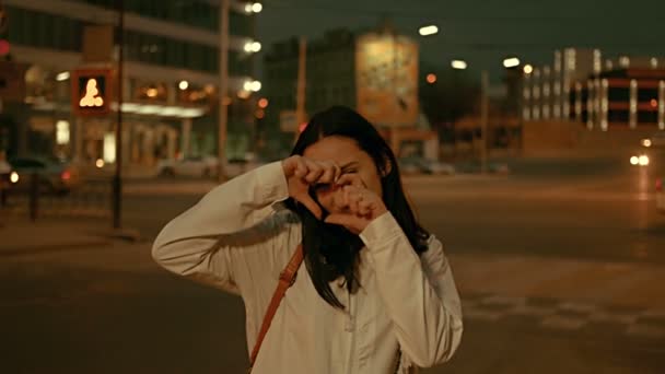 Joven mujer haciendo símbolo del corazón og sus dedos y ocultar su cara bonita en frente de la calle de la ciudad de noche con luces de coche desenfocadas y farolas — Vídeo de stock