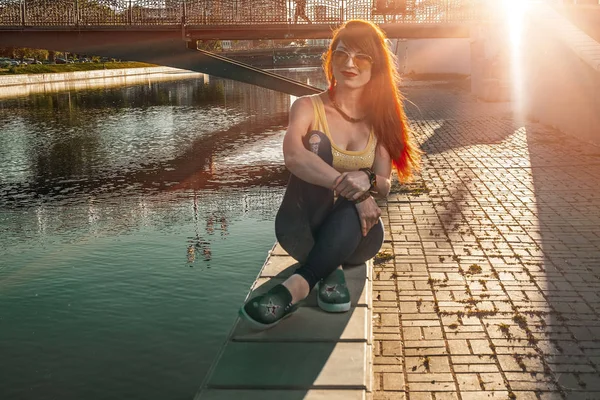 Gyömbér haj nő ül a város folyó töltésén háttérvilágítású napnyugta — Stock Fotó