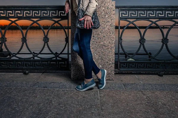 Look de moda callejera con jeans, zapatos de tenis y bolso de cuero negro. Niña posando en el terraplén del río frente al atardecer — Foto de Stock