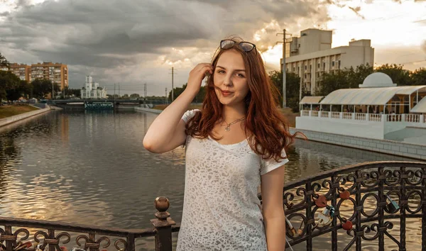 Gyömbér hajú nő a híd kerítés látszó-on fényképezőgép érint a haját előtt folyó és a felhős ég a szerelem zárak. — Stock Fotó