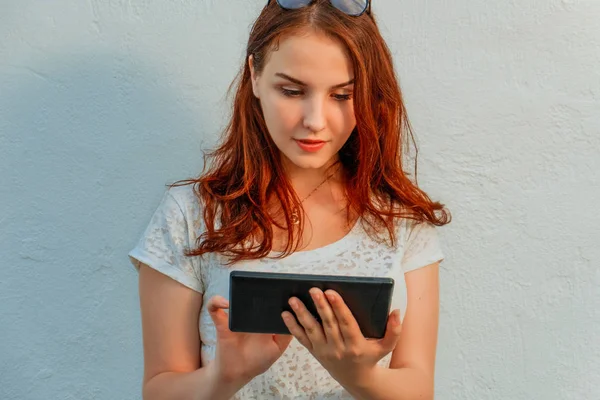 Элегантные рыжие волосы молодая деловая женщина или студентка с помощью планшета, опираясь на белую стену, глядя на экран — стоковое фото