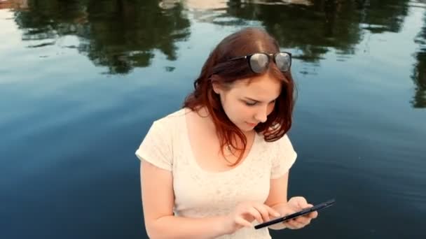 Mujer joven usando su tableta mientras se relaja al aire libre en un parque en un hermoso día de primavera cerca del río — Vídeos de Stock