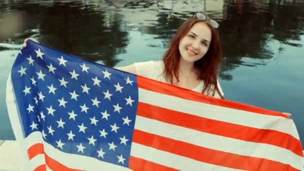 День независимости США, 4 июля. Красивая молодая девушка в случайном стиле улыбается с американским флагом в руках. Рыжие волосы, вода на заднем плане — стоковое видео