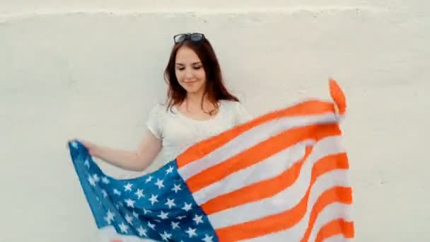 Привлекательная патриотичная молодая женщина держит американский флаг перед своим телом, танцуя перед стеной, празднуя июль — стоковое видео