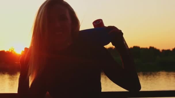 Hermosa rubia posando al atardecer junto al río sosteniendo un pequeño monopatín — Vídeo de stock