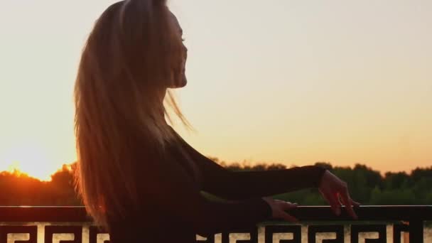 Hermosa rubia posando al atardecer junto al río parada junto a la valla y girando — Vídeo de stock