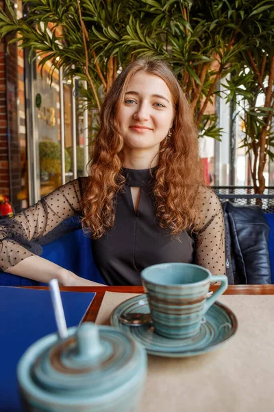 Ragazza riccia con i capelli rossi posa in un caffè. Sul tavolo davanti a lei ci sono tazze e zuccheriera . — Foto Stock