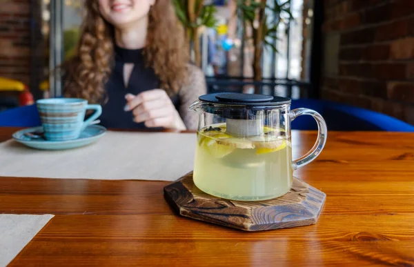 A kettle of tea made of herbs is on the table and a smiling girl sits in the background with a cup. — Stock Photo, Image