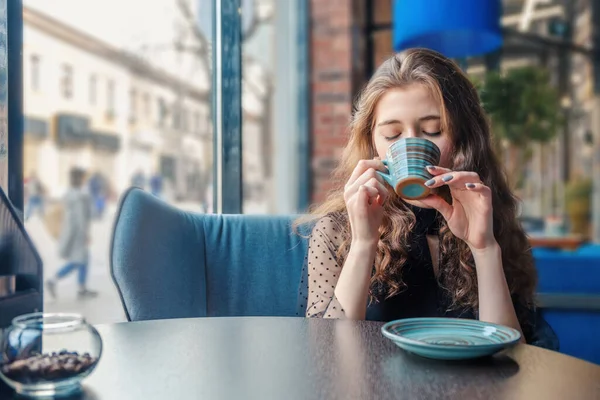 Een krullend meisje zit in een café bij een groot raam en drinkt uit een kopje dat haar ogen sluit. — Stockfoto