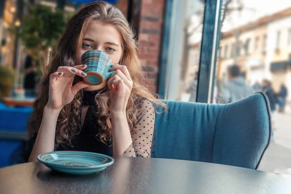 Roodharig meisje in een cafe drinkt een warm drankje uit een kopje en kijkt naar de camera — Stockfoto