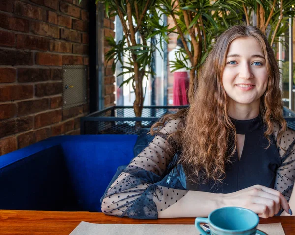 A smiling sly, rummaging girl looks directly into the camera sitting at a table in a cafe.