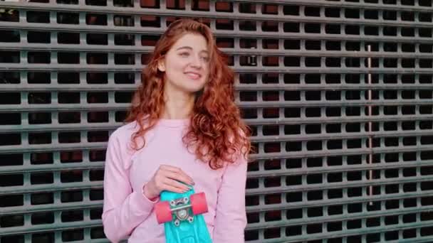 Een krullend roodharig meisje poseert tegen de achtergrond van een muur van metalen gaas in een skatepark, schieten met handen. — Stockvideo
