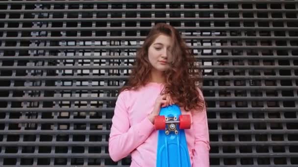 Una ragazza felice in un pullover rosa posa con uno skateboard tra le mani sullo sfondo di una griglia metallica . — Video Stock