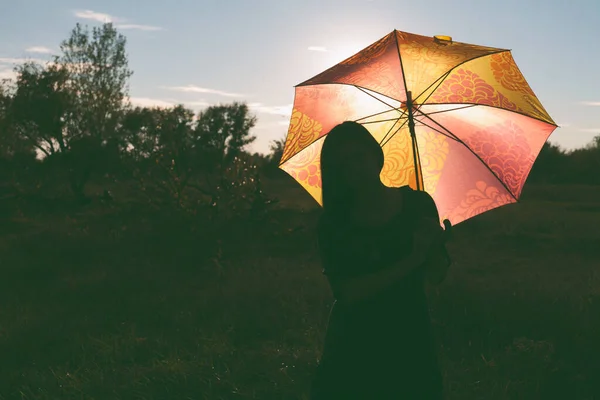 Una ragazza con un ombrello di colore posa in un campo. L'ombrello è illuminato da dietro dalla luce del tramonto — Foto Stock