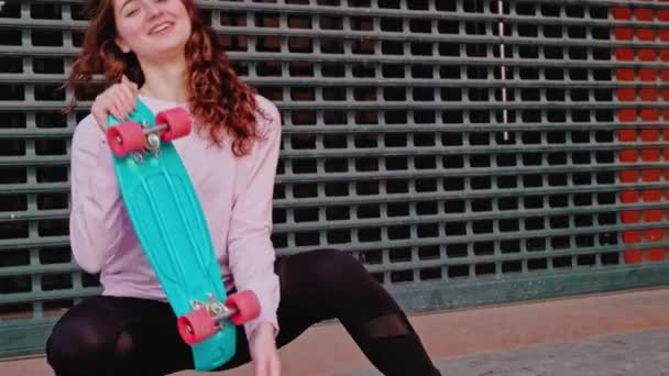 Uma menina ruiva posa com um skate em um parque de skate contra o pano de fundo de uma cerca de metal . — Vídeo de Stock