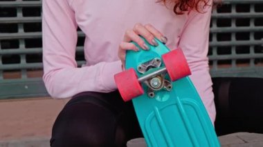 Spor kıyafetli genç bir kızıl saçlı mavi kaykayıyla oturmuş kameraya gülümsüyor. Biraz utangaç olması dışında keyfi yerinde..