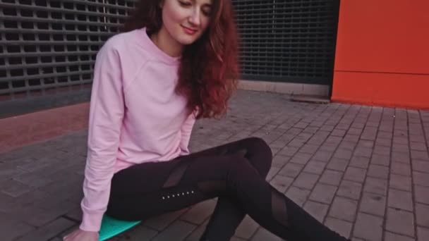 Una giovane ragazza con lunghi capelli rossi ricci siede su uno skateboard e sorride timidamente alla telecamera. La telecamera va da un piede all'altro. La clip è colorata di colore retrò . — Video Stock