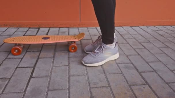 Menina fica ao lado de um skate laranja ao lado de uma parede brilhante . — Vídeo de Stock