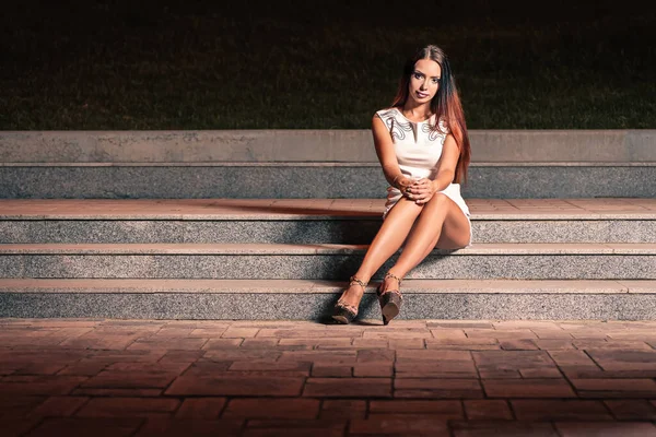 A girl in a short dress with long bare feet sat down to rest on the steps at night. She looks straight into the camera where her long legs are stretched.