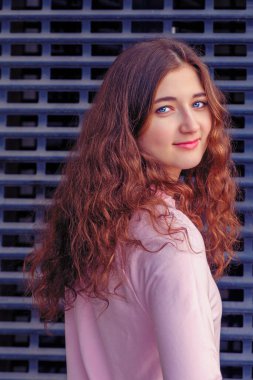 Uzun kıvırcık kahverengi saçlı kız kameraya döner ve önüne bakar. Demir bir ızgaranın önünde duruyor..