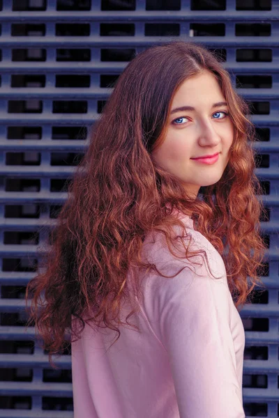 La fille aux longs cheveux bruns bouclés se tourne vers la caméra et regarde droit devant elle. Elle se tient devant une grille de fer . — Photo