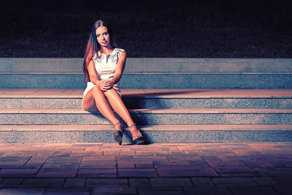 Mujer en un vestido corto con los pies descalzos largos se sentó a descansar en las escaleras por la noche. Ella mira directamente a la cámara donde sus largas piernas están estiradas . — Foto de Stock