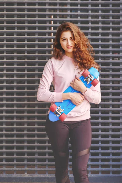 Porträtt av en flicka med en skateboard mot bakgrund av en metall skyddsnät i en skateboard park. Hon trycker skateboarden mot bröstet och ler blygt mot kameran. — Stockfoto