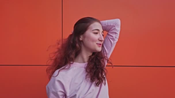 Una jovencita contra una pared roja cepilla su largo cabello rojo con sus dedos y luego mira a la cámara con una amplia sonrisa — Vídeos de Stock