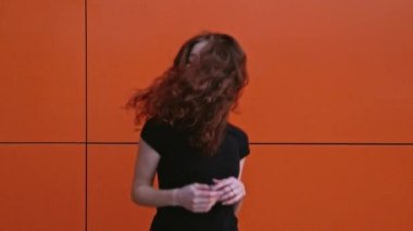 Kızıl saçlı kız spor kıyafetleriyle kırmızı duvara karşı ağır çekimde dans ediyor.