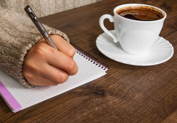 Woman's hand in sweater writing on notepad at desk. cup of coffee on the wooden table. — Stock Photo, Image