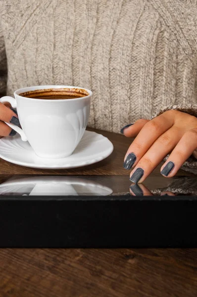 Manos de mujer en suéter sosteniendo la taza de café y utiliza un dispositivo de tableta en blanco en la mesa de madera . — Foto de Stock