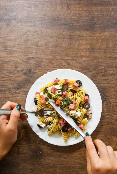 woman hand holding fork and knife with pasta fusilli salad with salami, cheese and olive in white plate on a wooden background. with copy space. top view