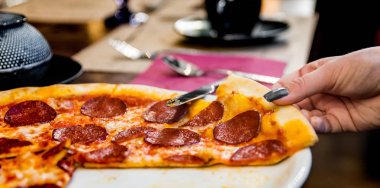 Kadın El, Mozzarella peyniri, salam, domates, biber ve baharatlı Pepperoni Pizza alır. Kafedeki ahşap masa arkasında İtalyan pizzası