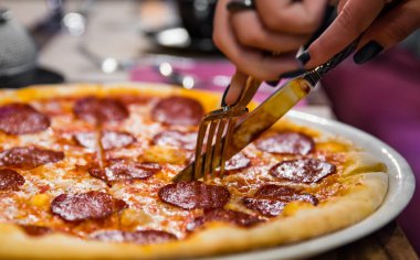Kadın eli bıçaklı ve çatallı kahve masasında pepperonili pizza kesiyor.