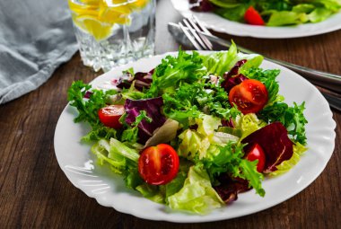 Tahta arka planda beyaz tabakta domatesli çeşitli taze salata yaprakları..