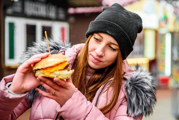 ストリートフード ジューシーなバーガーを持って冬の外で食べる若い女性 — ストック写真
