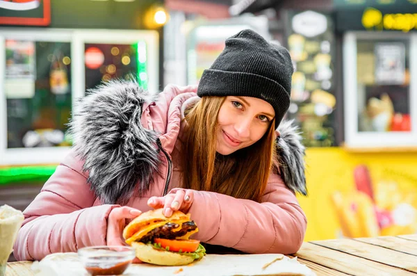 ストリートフード ジューシーなバーガーを持って冬の外で食べる若い女性 — ストック写真