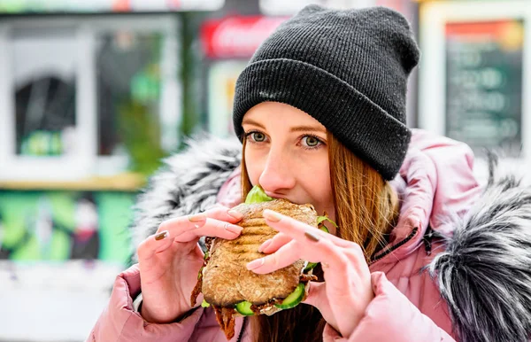 ストリートフード ジューシーなシアバタサンドイッチを持って冬の屋外で食べる若い女性 — ストック写真