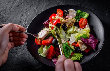 Bıçak ve çatallı kadın elleri tavuk göğsü, peynir, siyah zeytin, kırmızı biber, marul, taze salata ve siyah bir tabakta domates yer.