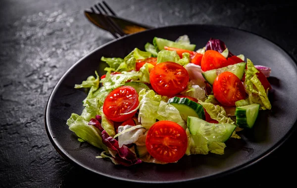 various fresh mix salad leaves with tomato, cucumber and red pepper on a black plate on Dark grey black slate background