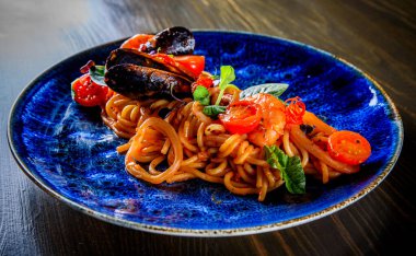 İtalyan spagetti makarna marinara, deniz tarağı, karides, midye, domates, ahşap masa arkasında mavi tabakta deniz ürünleri kokteyli.