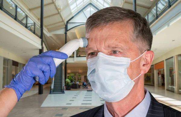 Senior mand iført ansigtsmaske med temperatur taget for at kontrollere for virus i butikker - Stock-foto