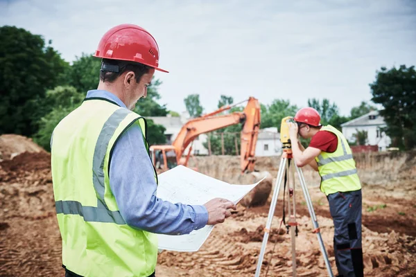 Trabajadores de la construcción en la zona del edificio. Capataz con plano y agrimensor — Foto de Stock