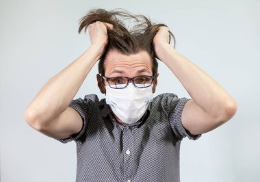 Tıbbi maske takan genç bir adam ellerini gri bir arka planda tutuyor. Koronavirüs salgını panik duyguları kavramı.