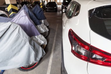 Motosikletler ve beyaz araba kapalı bir yeraltı otoparkında park halindedir.