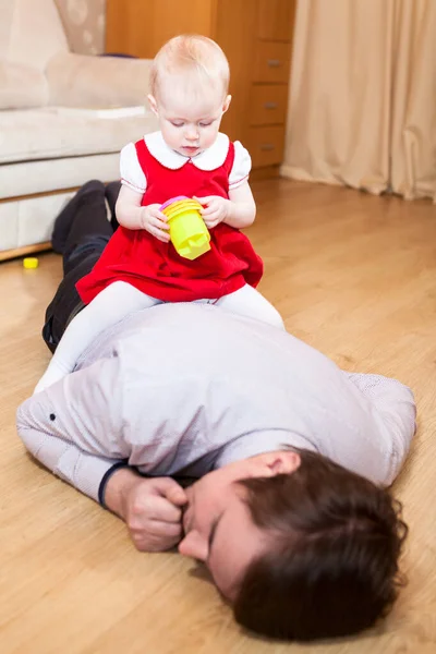 小女儿坐在疲惫的父亲的背上 昏迷不醒地躺在地板上 男人在和孩子玩玩具后睡着了 — 图库照片