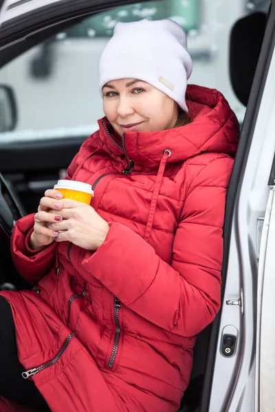 魅力的な大人の女性ホットコーヒーと紙のガラスを保持し 冬にドアを開けて車の中で座って — ストック写真
