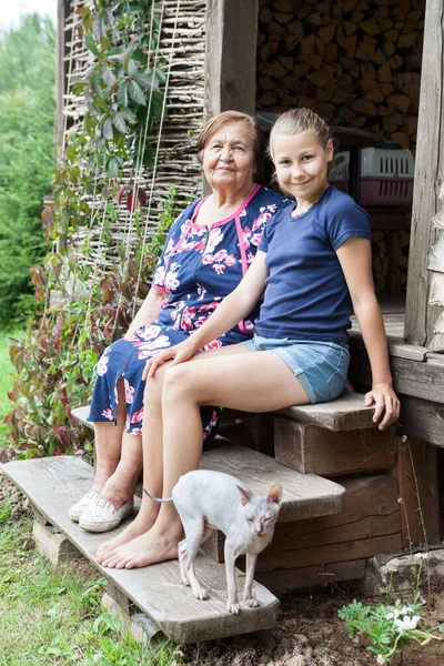 年迈的白人妇女 带着孙女和斯芬克斯猫坐在村舍门廊的木楼梯上 夏季来临 — 图库照片