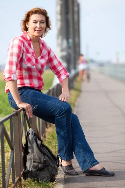 穿着红色T恤 头戴蓝色牛仔裤 背着背包坐在街上的成熟白人妇女的画像 — 图库照片