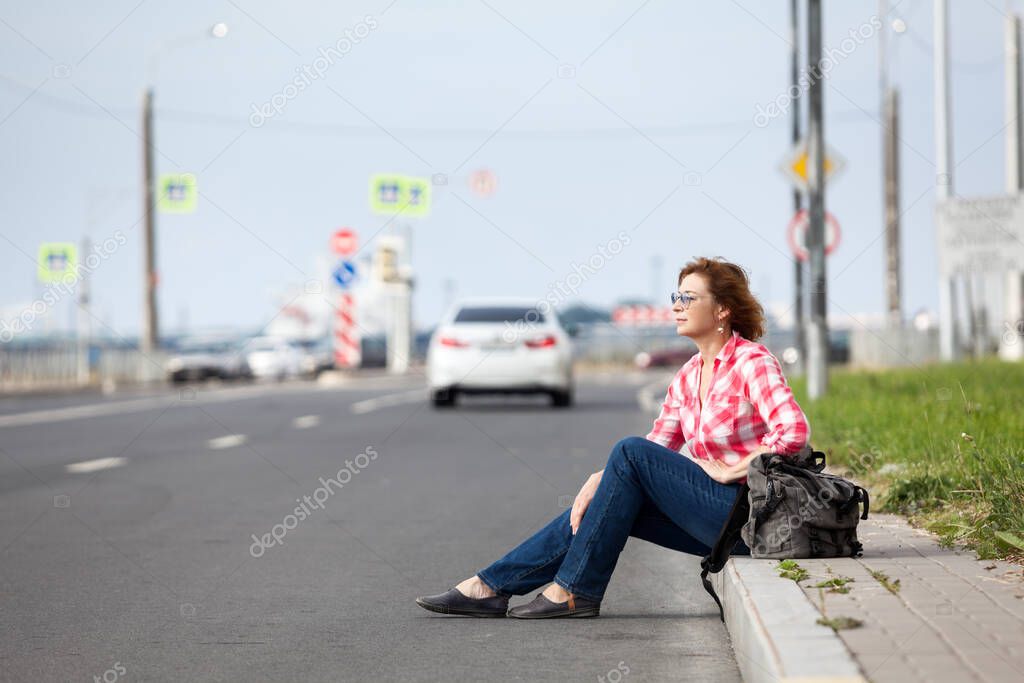 Hitchhiking, passenger car driving away, woman sitting roadside with backpack